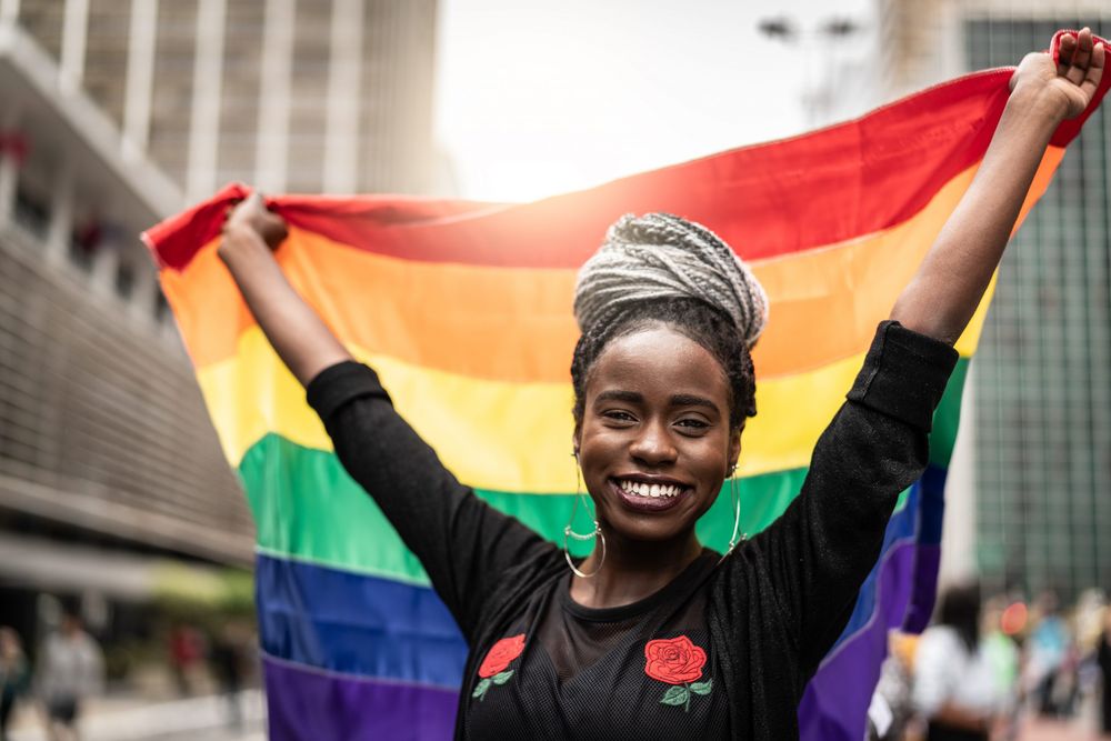 Smiling Black woman with wrapped hair holding a rainbow LGBTQ+ pride flag