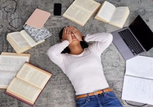 Black woman laying on floor with hands over face surrounded by open books and laptop
