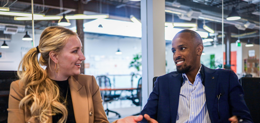 A White woman with long blond hair speaking to a Black man with a goatee and short brown hair in an office setting
