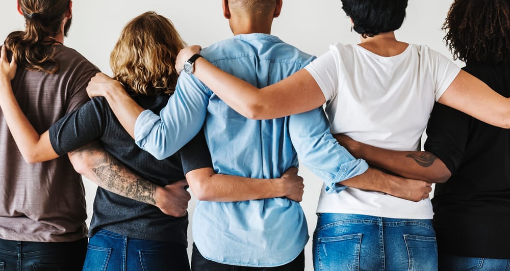 Five people of different ethnicities stand with their backs to the camera and arms entwined.