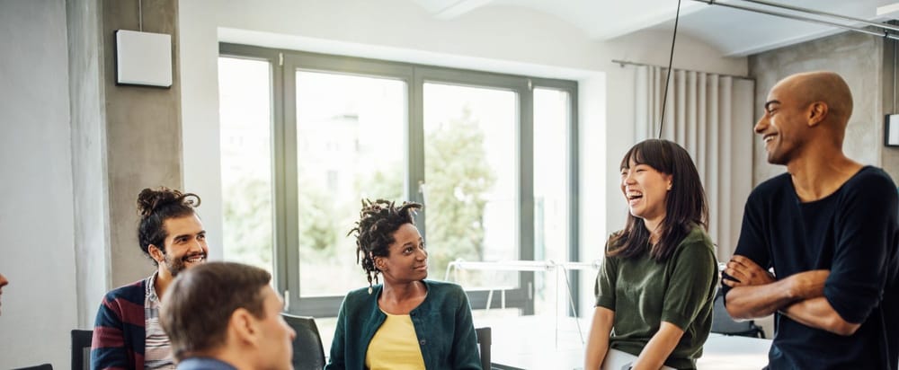 Colleagues looking at cheerful businesswoman holding laptop in meeting at creative office