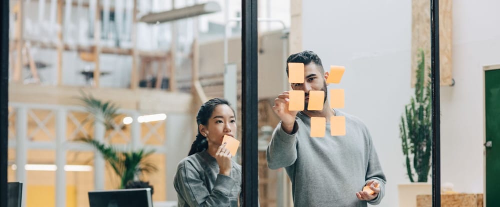 Colleagues discussing while sticking adhesive notes on glass wall in office