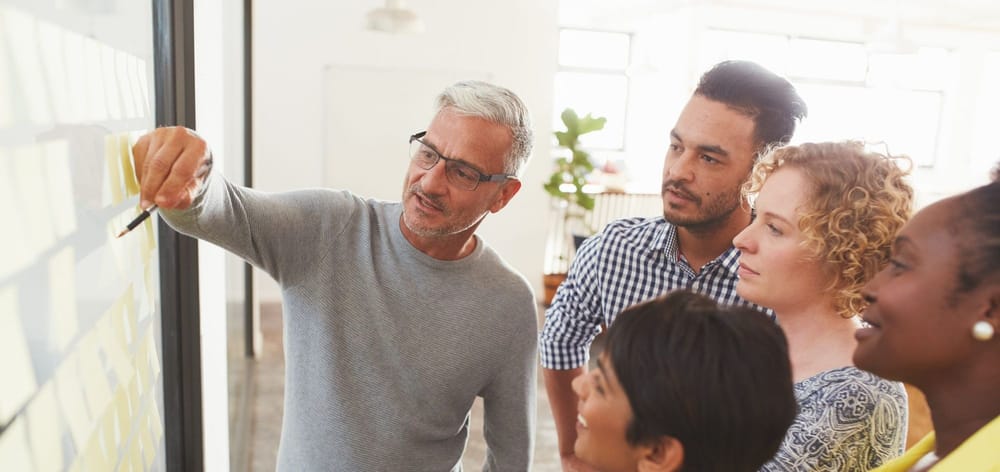 Mature businessman and his diverse team brainstorming with yellow adhesive notes on a glass wall in a bright modern office